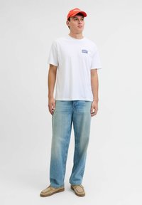 Jeune homme portant une casquette rouge, un t-shirt graphique blanc, un jean bleu clair et des chaussures décontractées beiges, se tenant contre un fond blanc uni.