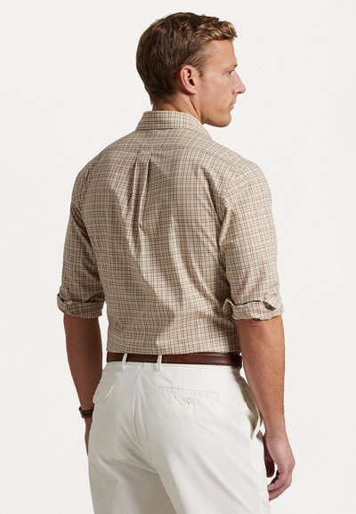 Chemise à manches longues en tartan aux tons beiges et bruns avec des manches retroussées. Le dos présente un pli central et un ourlet arrondi. Portée avec un pantalon clair.