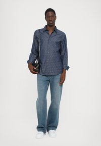 Camisa abotonada azul marino con patrones blancos, combinada con jeans de mezclilla claros de pierna acampanada. El modelo sostiene un pequeño bolso cruzado negro. Zapatillas blancas.