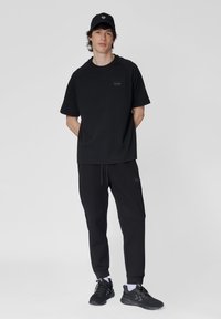 Schwarzes Baumwoll-T-Shirt und passende Jogginghosen, mit Rundhalsausschnitt, lässiger Passform und Logo-Akzenten. Kombiniert mit schwarzen Sneakers und einer Mütze.