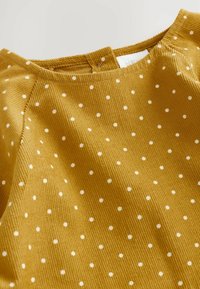 Top giallo senape realizzato in tessuto testurizzato con pois bianchi. Presenta un collo rotondo e una chiusura sul retro per facilità di indossamento.