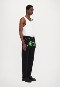 Black denim pants with green cat graphic on the front, paired with a white tank top and black lace-up boots. Simple, straight-leg design.