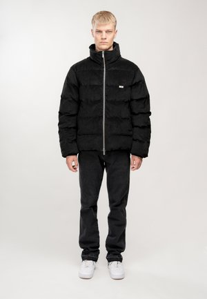 Schwarze Daunenjacke mit hohem Kragen, horizontal gesteppten Abschnitten, vorderem Reißverschluss und kleinem Logo auf der linken Brust. Kombiniert mit schwarzen Hosen.
