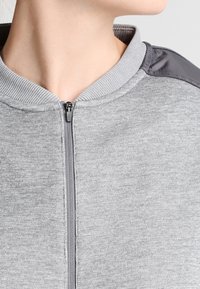 Giacca grigia con zip e colletto a coste, caratterizzata da un tessuto grigio liscio sulle spalle e un corpo testurizzato. Dettaglio della zip incluso.