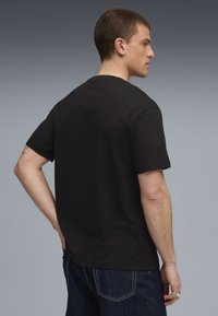 Mand iført ensfarvet sort t-shirt og mørkeblå jeans, der står med siden til med den ene hånd på hoften mod en grå gradient baggrund.