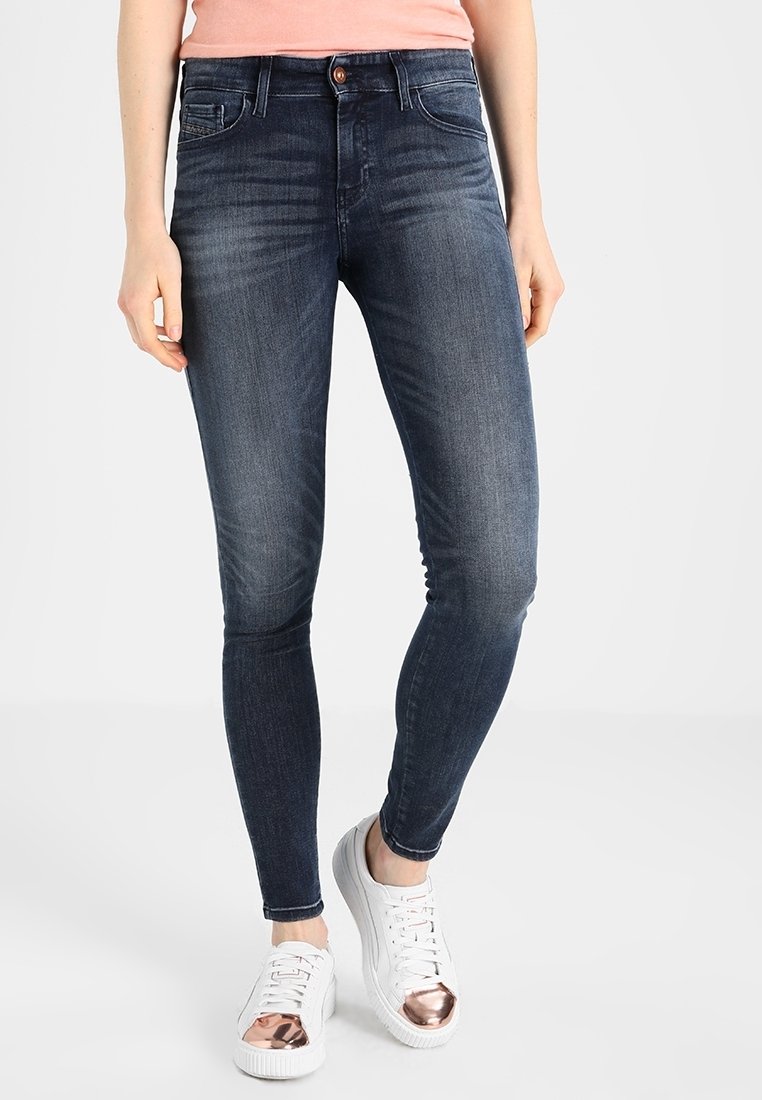 Jeans ajustados de denim azul oscuro con un lavado desgastado, diseño de cinco bolsillos y botón dorado. Combinados con zapatillas blancas con detalles en oro rosa.