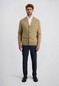 Mann steht frontal und trägt eine beigefarbene, geknöpfte Strickjacke über einem weißen Hemd, marineblaue Hose und dunkelbraune Slipper vor einem schlichten Hintergrund.