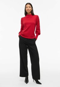 Pull en tricot rouge avec des manches texturées, associé à un pantalon large noir et des bottes à talons pointus. Tissu lisse et design ajusté.