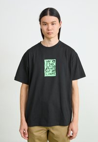 T-shirt en coton noir avec un graphique vert vif représentant un personnage et le mot "FLEUR". Manches courtes et encolure ras du cou.