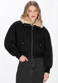 Schwarze, kurz geschnittene Jacke mit einem beigen Kunstfellkragen, Reißverschluss vorne, zwei Seitentaschen und gerippten Bündchen. Glattes Material mit strukturiertem Schnitt.