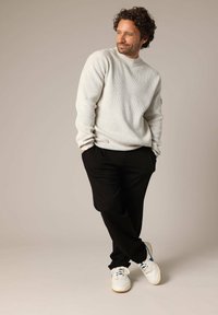 Lichtgrijze ribbeltrui, zwarte broek en witte sneakers met blauwe accenten. Het model staat casual met de handen in de zakken.