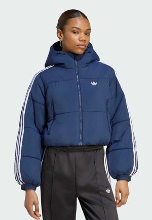 Jonge vrouw die een blauwe donsjas draagt met witte strepen op de mouwen en zwarte Adidas broek met een wit logo en strepen.