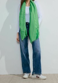 Femme portant une écharpe verte vif sur un top blanc à manches longues, un jean bleu à jambes larges et des baskets blanches avec des touches rouges.