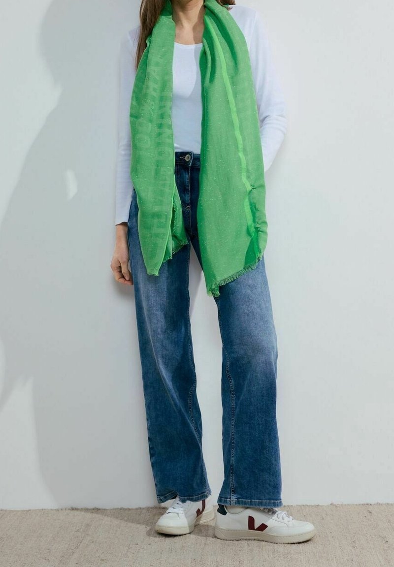 Femme portant une écharpe verte vif sur un top blanc à manches longues, un jean bleu à jambes larges et des baskets blanches avec des touches rouges.