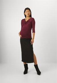 Maternity outfit featuring a deep burgundy fitted top with a twist detail, paired with a black ankle-length skirt and black heeled boots.