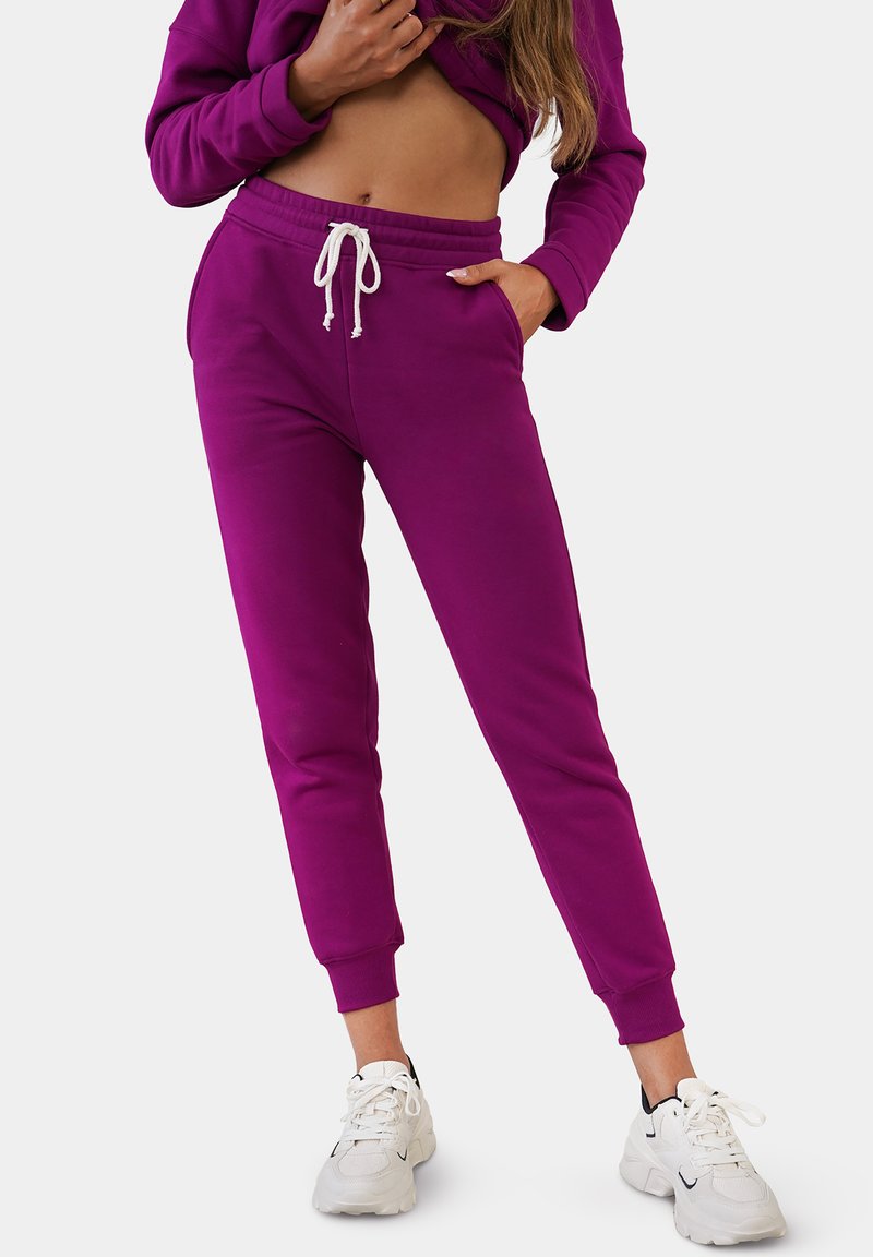 Lila Jogginghose mit elastischem Bund und Kordelzug, mit schmal zulaufenden Beinen und Bündchen an den Knöcheln. Trägt weiße Sneaker.