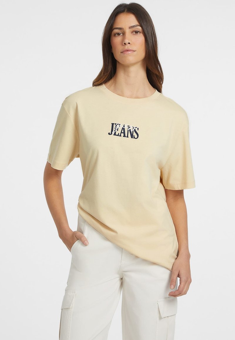 Koszulka z krótkim rękawem w jasnym beżowym kolorze, wykonana z bawełny, z granatowym napisem "JEANS" na środku. Luźny krój i okrągły dekolt.