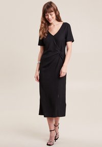 Schwarzes Kurzarmkleid mit Wickeldesign, strukturiertem Stoff und einem seitlichen Schlitz. Kombiniert mit schwarzen Riemchenschuhen und einem silbernen Armband.