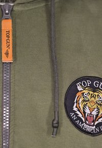 Olivengroene hoodie met ritssluiting, voorzien van een leren label met de tekst "TOP GUN" en een ronde geborduurde tijgerpatch.
