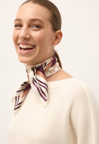Une écharpe en soie présente des motifs géométriques en crème, bordeaux et mauve, nouée soigneusement autour du cou, avec des extrémités pointues et une texture lisse.