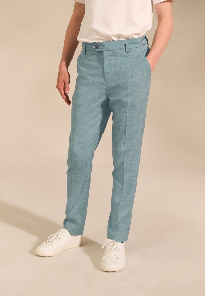 Pantalon bleu clair en tissu texturé, avec poches avant, devant plat et passants pour ceinture. Porté avec des baskets blanches.