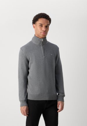 Uomo che indossa un maglione grigio con mezza cerniera, collo, polsini e orlo a coste, abbinato a pantaloni neri, su uno sfondo chiaro e uniforme.