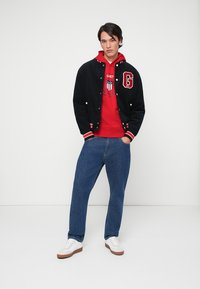 Veste varsity noire avec des accents rouges, associée à un sweat à capuche rouge avec un logo circulaire, un jean bleu et des baskets blanches avec des semelles brunes.