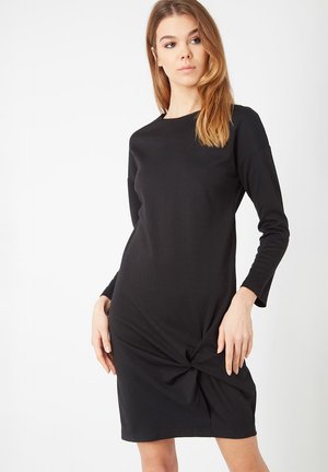Vestido de punto negro con mangas largas, escote redondo y un detalle de nudo torcido en la cintura. Textura suave y silueta ajustada.