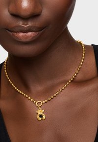 Collana di perle d'oro con un fermaglio rotondo, dotata di un ciondolo con disegni traforati e un accento di pietra scura, visibile su un modello dalla pelle scura.