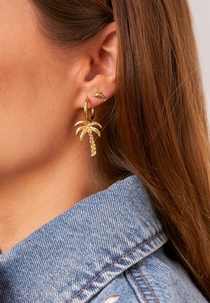 Ohr mit zwei goldenen Ohrringen: einem kleinen Delfin-Stecker und einem Creolenring mit Palmenanhänger, braune Haare und sichtbarer Kragen einer blauen Jeansjacke.