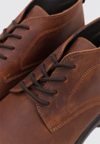 Zapatos de cuero marrón con punta redondeada, costuras tono sobre tono y cordones marrón oscuro. Detallados con un acabado liso y costuras laterales sutiles.