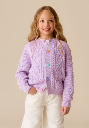 Ung pige med langt blondt hår iført en lavendel kabelstrikket cardigan med flerfarvede knapper og hvide bukser, smilende og kiggende op.