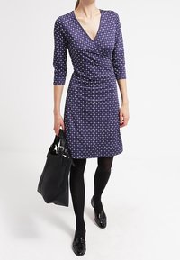 Námořnické zavinovací šaty s bílými puntíky, tříčtvrteční rukávy, po kolena, doplněné černými punčochami a lesklými černými botami, nese černou kabelku.