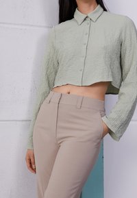 Camisa corta de color verde claro, texturizada, con botones en la parte delantera y mangas largas, combinada con pantalones beige ajustados.
