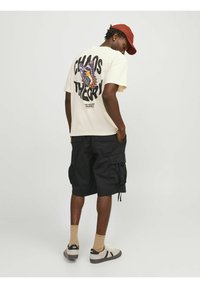 Cremes T-Shirt mit dem Text "CHAOS THEORY" und einem Schmetterlingsgrafik auf dem Rücken, kombiniert mit schwarzen Cargo-Shorts, beigen Socken und Freizeitschuhen.