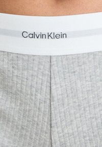 Sivkasta rebraste tkanine z belim pasom, na katerem je v črni pisavi napis "Calvin Klein". Pas ima subtilno sivo črto.