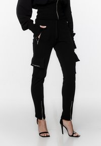 Czarne spodnie cargo z bocznymi kieszeniami i zamkami błyskawicznymi u dołu, wykonane z miękkiego materiału. Posiadają widoczną łatkę z logo na kieszeni.