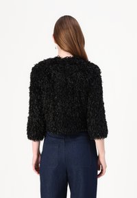 Svart kort jacka med en texturerad, fluffig yta och trekvartsärmar, tillsammans med högmidjade mörka jeans.