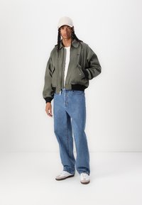 Casaco bomber verde-azeitona com fecho na frente, punhos canelados pretos, combinado com jeans largos azuis claros e ténis brancos. Um boné bege completa o look.