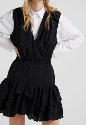 Kobieta w czarnej, warstwowej sukience bez rękawów na białej koszuli z kołnierzykiem, kolorowe kolczyki z koralików, ręce na biodrach, jasnoszare tło.