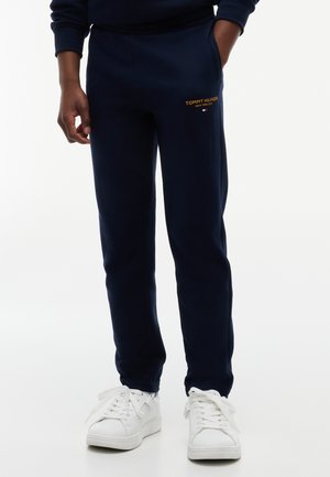 Person trägt dunkelblaue Tommy Hilfiger Jogginghose und weiße Sneakers mit den Händen in den Taschen vor weißem Hintergrund.