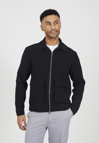 Giacca nera con zip, caratterizzata da un colletto, due tasche frontali, una texture liscia e un design aderente. Indossata sopra una camicia bianca.