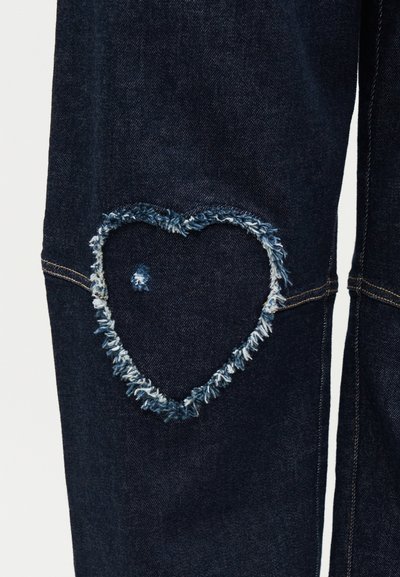 Σκούρο μπλε τζιν με σκισμένη καρδιοειδή επιδιόρθωση στην περιοχή του γόνατου και ορατή πορτοκαλί ραφή κατά μήκος των ραφών.
