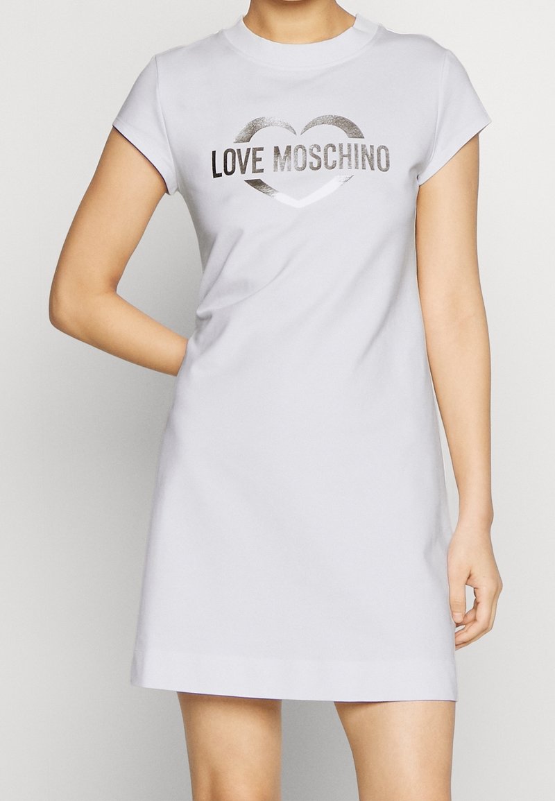 Personne portant une robe blanche à manches courtes avec le texte argenté "LOVE MOSCHINO" et un contour de cœur sur la poitrine, debout.
