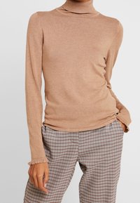 Pull à col roulé marron clair en tissu doux, avec des manches longues à poignets volantés, associé à un pantalon à motif à carreaux.