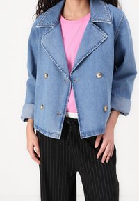 Chaqueta de mezclilla en azul claro con un diseño de doble botonadura, grandes solapas, dos botones y mangas enrolladas. Combinada con una camisa rosa y pantalones a rayas.