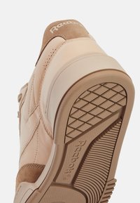 Beige sneaker with suede and leather materials, featuring embossed logo, textured rubber sole, and subtle perforations for ventilation.