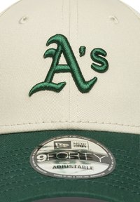 Gorra de béisbol ajustable que presenta una parte superior crema y un visor verde. En la parte frontal aparece un logo bordado en verde con la letra "A's". La etiqueta dice "New Era 9FORTY".