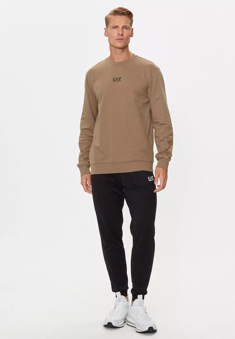 Felpa marrone con logo, abbinata a pantaloni jogger neri. Il modello indossa sneakers bianche. Design semplice, vestibilità casual, materiale in misto cotone.