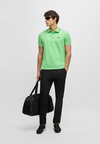 Grön polo med en svart logotyp, svart trim på kragen och ärmarna, kombinerad med svarta byxor och en svart sportväska.